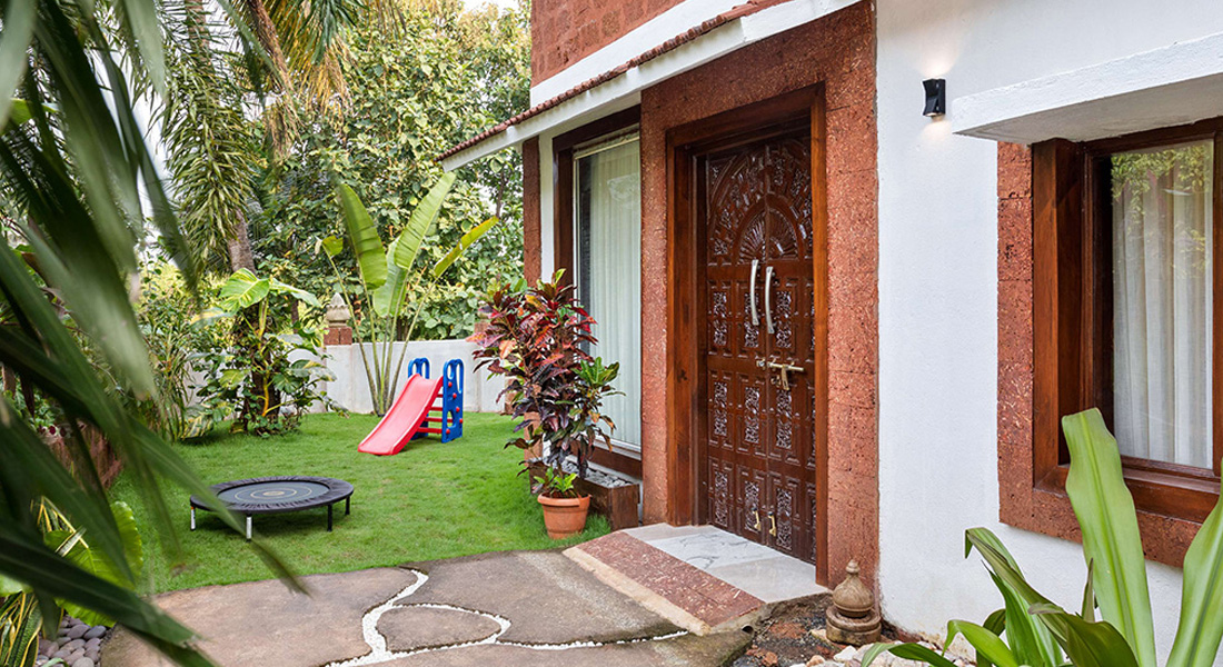 Villa exterior with private garden, green lawn, wooden entrance door, tropical plants, and a children’s play slide