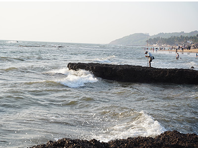 Rocky shoreline at Anjuna Beach with waves crashing along the coast and visitors enjoying the beach in North Goa