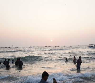 People enjoying sea waves and sunset views at Calangute Beach in Goa with boats in the background.