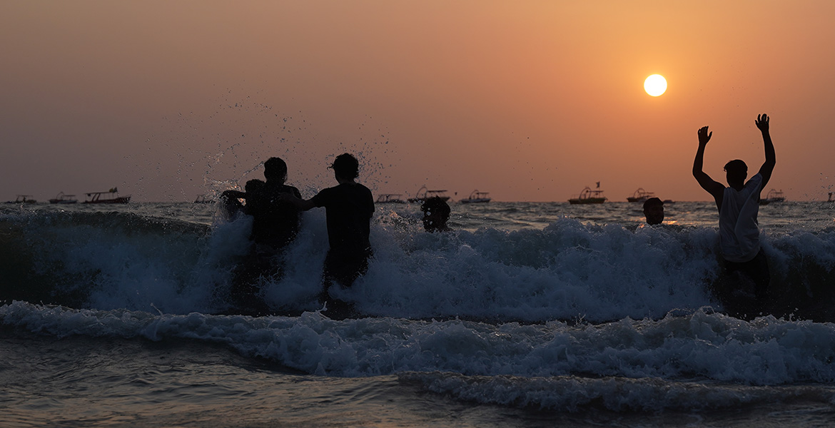 People enjoying sea waves during sunset at Calangute Beach with silhouettes, ocean waves, and boats in the background