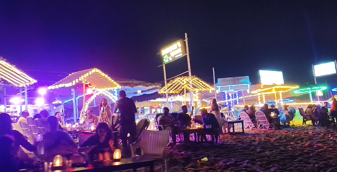 Colorful beachside nightlife with illuminated shacks, dining tables, and tourists at Candolim Beach in Goa