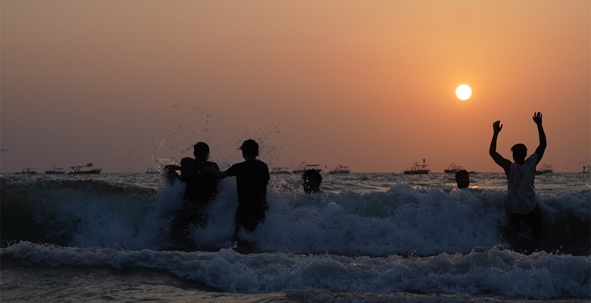 People swimming in the sea at sunset on a Goa beach with waves, fishing boats and an orange sky