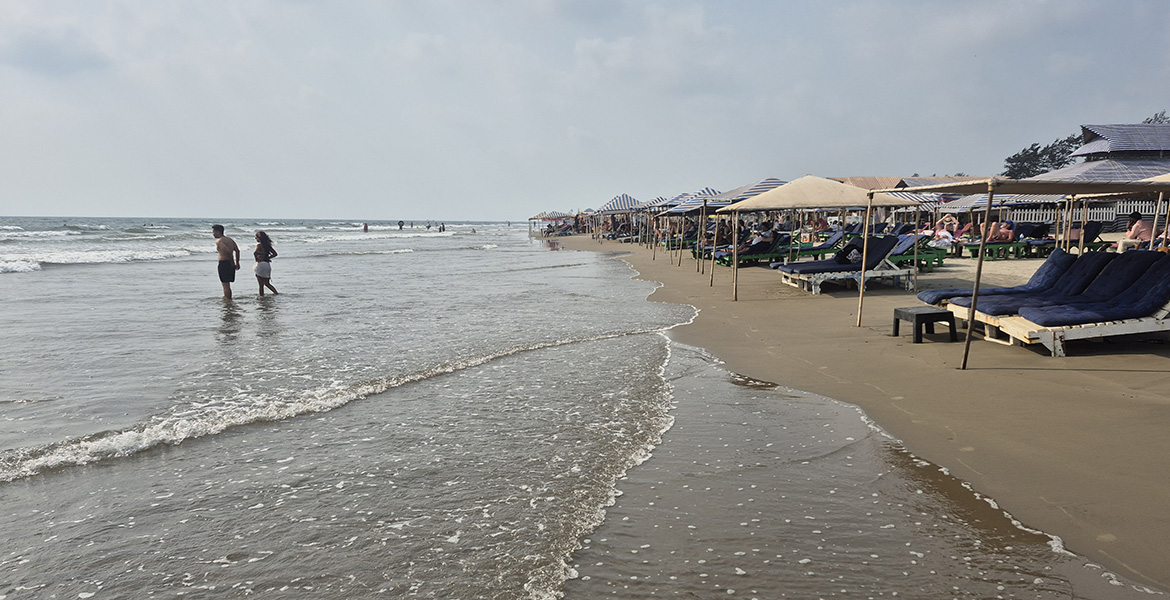 Visitors walking along the shoreline at Morjim Beach in Goa with beach shacks and lounge seating beside the sea