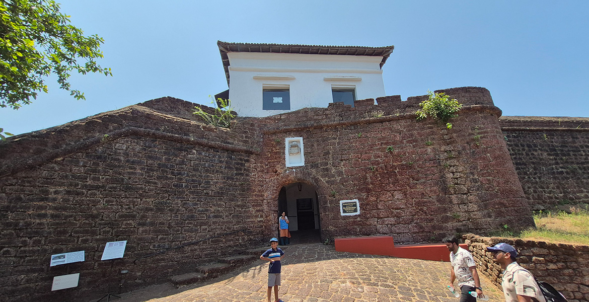 Main entrance of Reis Magos Fort in Goa with historic stone walls, arched gateway, and visitors standing outside under a clear blue sky