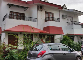 Modern Goan villa with white exterior, red roofing, balconies, and parked cars in front
