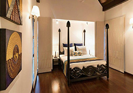 A beautifully designed luxury bedroom featuring a carved wooden four-poster bed, warm lighting, hardwood flooring, and decorative wall art.