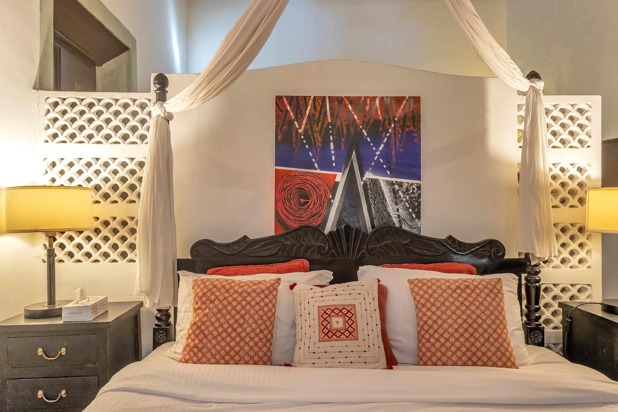 Four-poster bed with a dark wooden headboard, red/orange patterned pillows, and sheer canopy