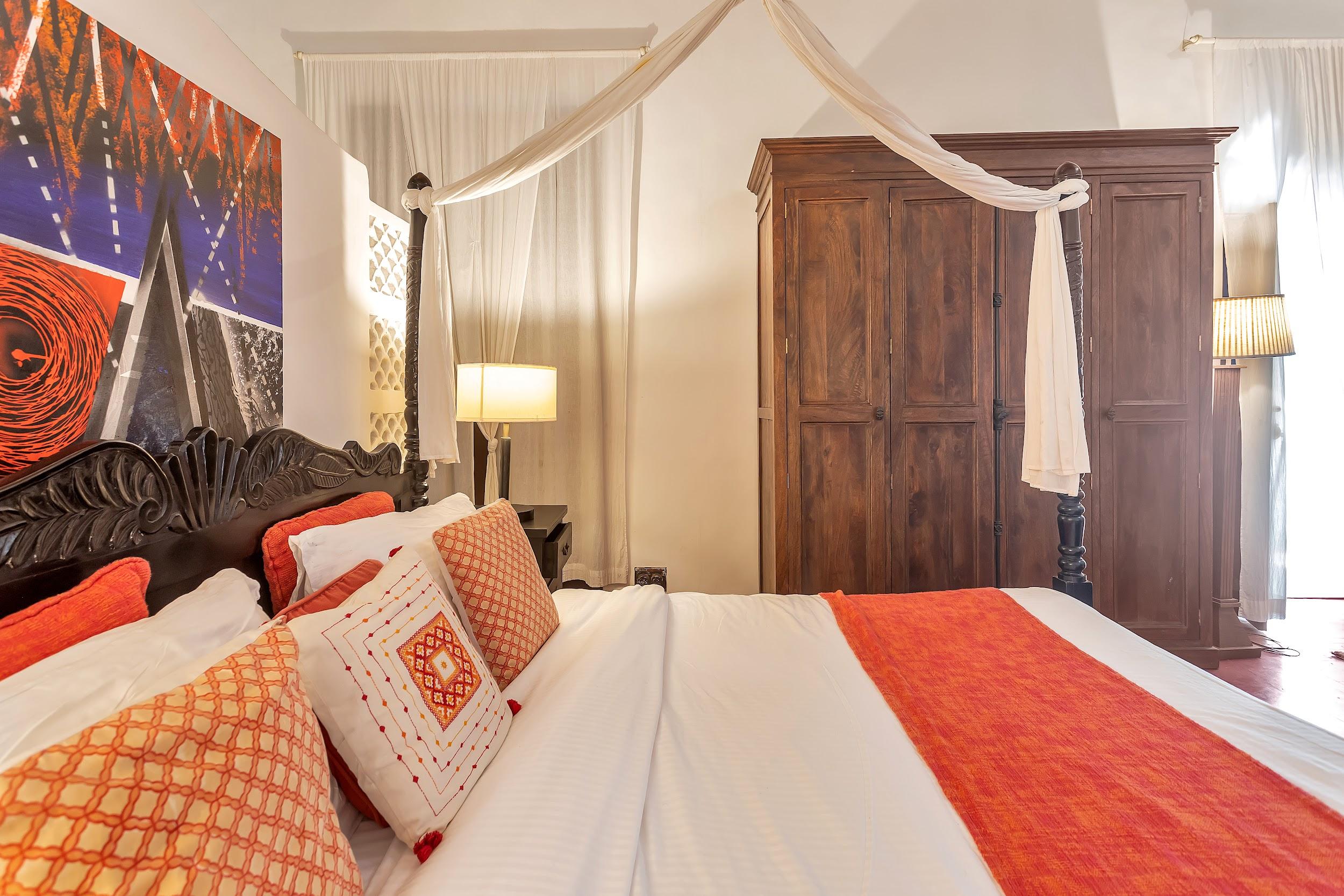 Close-up of a dark wood four-poster bed with bright orange accent pillows and a large wooden wardrobe
