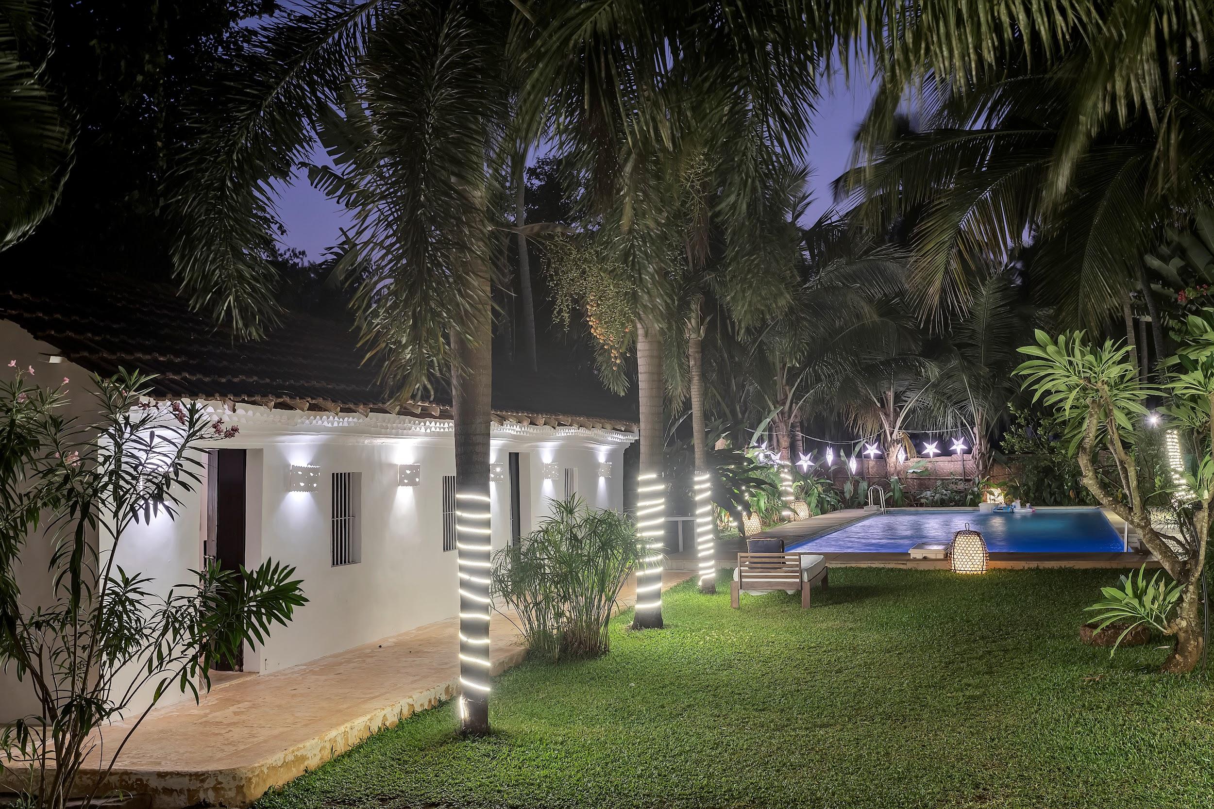 Exterior view of the villa at night, showing the pool area illuminated with string lights wrapped around palm trees