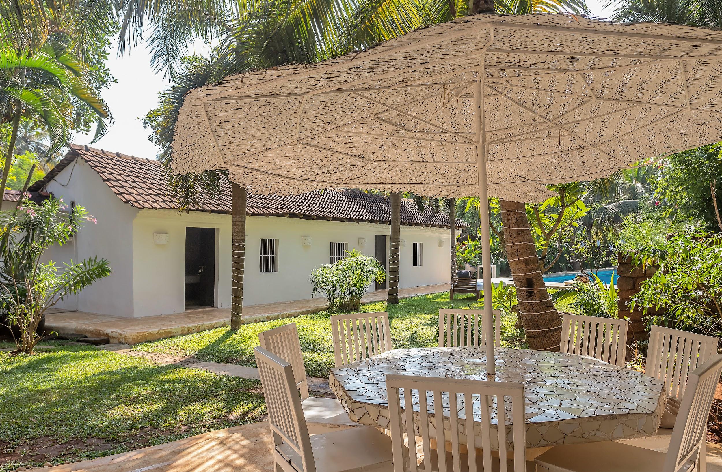 Outdoor dining set with a mosaic table and white chairs under a large thatched umbrella.