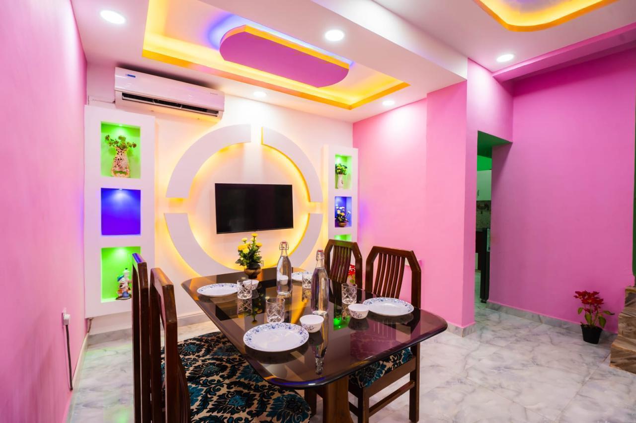 Dining area with glass table, wooden chairs, TV on decorative white wall, pink walls.